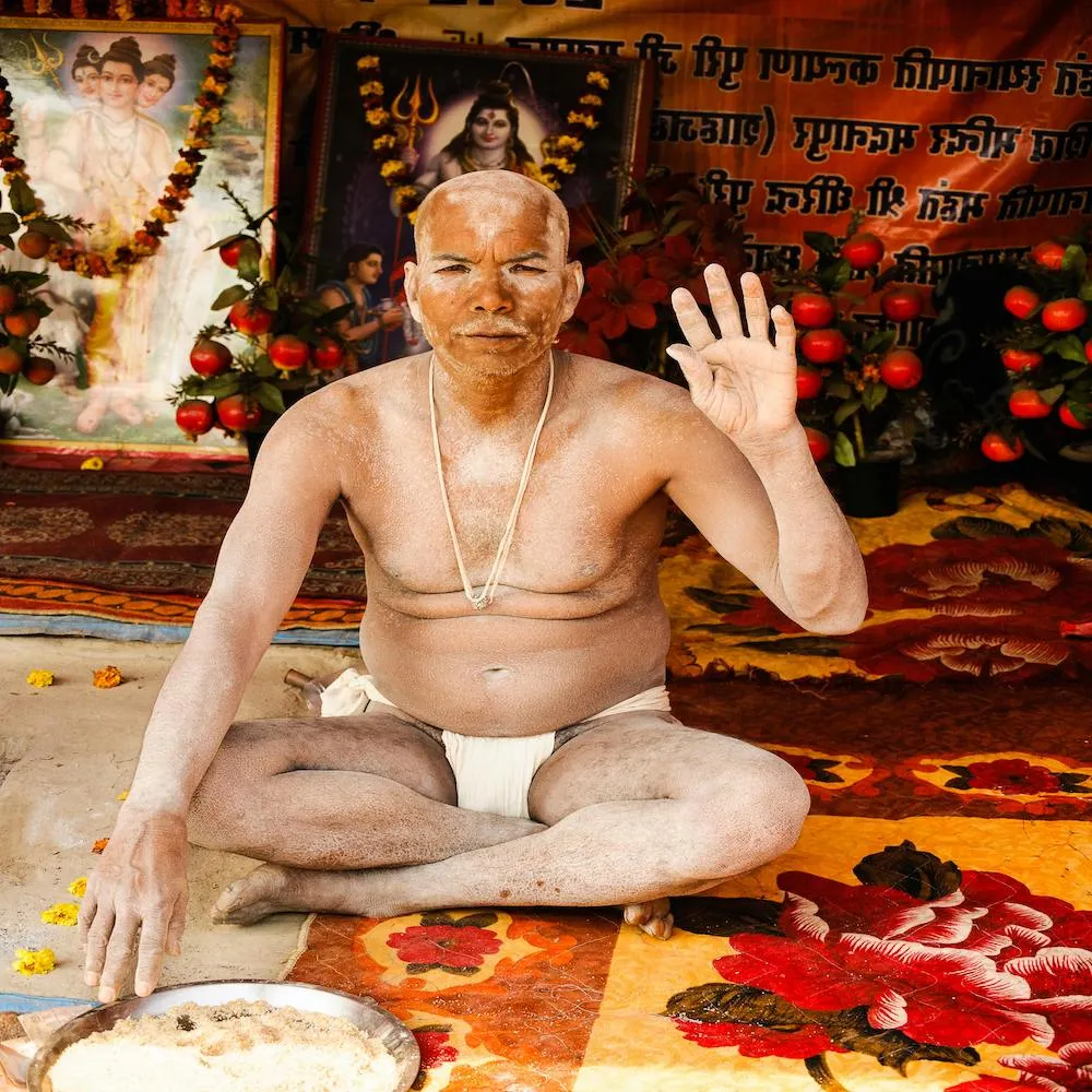 Indian man sitting cross-legged with mud or cow dung on his face and body, wearing traditional Hindu underwear resembling sumo-style wear.